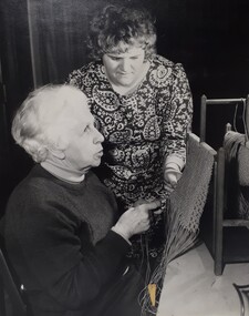 Elderly woman holds the threads, whilst another woman looks at the weaving the other has done.