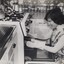 Woman wearing a pinafore and oven gloves kneels before an oven, about to remove a casserole dish