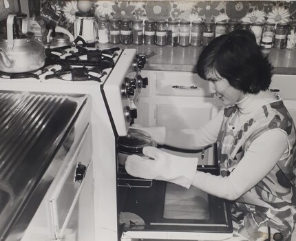 Woman wearing a pinafore and oven gloves kneels before an oven, about to remove a casserole dish