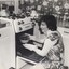 Woman wearing a pinafore and oven gloves kneels before an oven, about to remove a casserole dish