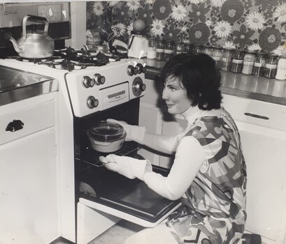 Woman wearing a pinafore and oven gloves kneels before an oven, about to remove a casserole dish