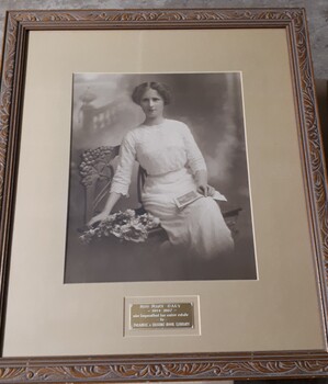 Sepia photograph of woman in early 20th century dress on bench next to flowers and with open book in her lap