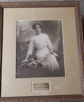 Sepia photograph of woman in early 20th century dress on bench next to flowers and with open book in her lap