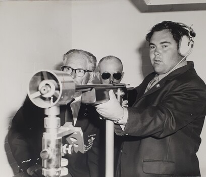 Man wearing ear muffs holding rifle in frame with two other men beside him
