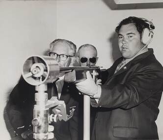 Man wearing ear muffs holding rifle in frame with two other men beside him