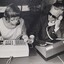 Woman touches device to a key on a switchboard, which a man to her left has pointed out as he holds a receiver to his ear