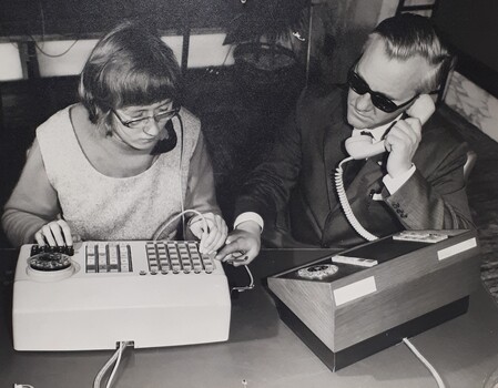 Woman touches device to a key on a switchboard, which a man to her left has pointed out as he holds a receiver to his ear
