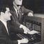 Man sits in front of a telephone switchboard, his hand holding a jack to plug into the socket, is guided by another man who stands