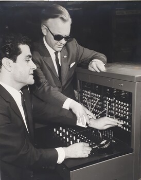 Man sits in front of a telephone switchboard, his hand holding a jack to plug into the socket, is guided by another man who stands
