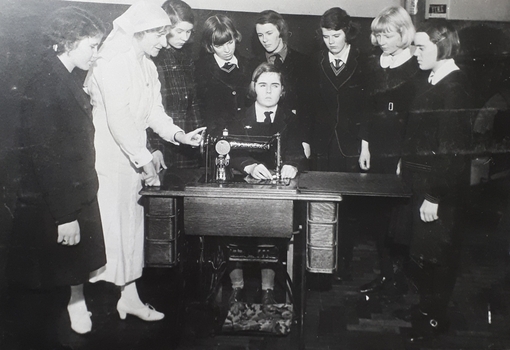 Schoolgirls and Sister huddle old treadle sewing machine watching a girl sew