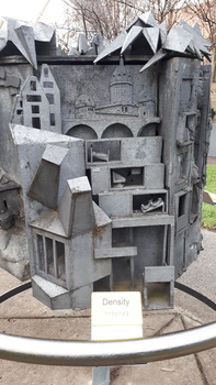 Multiple boxes, some with figures inside, are crowded beneath a bridge and dome.  A white and black sign reads 'Density' in writing and Braille