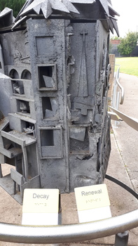 Boxes with broken doors and unfinished concrete depict 'Decay' and a church with a Calvary cross over roughened surface is 'Renewal'.  Both have white and black signs with writing and Braille.