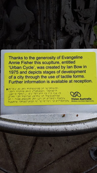 Yellow and blue sign providing name of artist, sculpture and person who commissioned it in writing and Braille