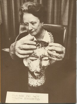 A seated woman touches a sculpture of head, before which rests a sign 'Please do not touch exhibits'