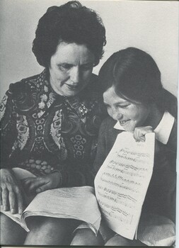 Woman reading Braille music score in her lap whilst a schoolgirl holding a printed score looks on