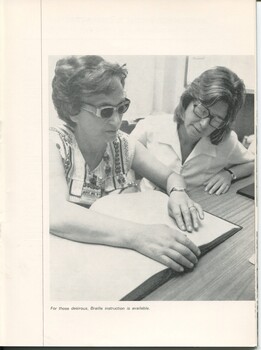 Woman sits at a table reading Braille whilst another looks on