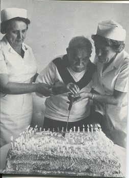 Man assisted by two nurses to cut cake celebrating his centenary birthday