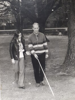 A woman walks beside a man who uses a white cane whilst crossing the park