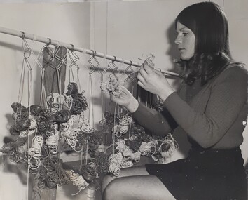 Lady in mini skirt sits as she weaves plastic cording into a pattern on a curtain rail