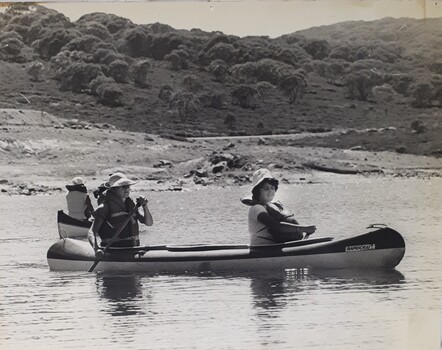 Two woman paddle a canoe across a lake whilst another canoe passes behind them