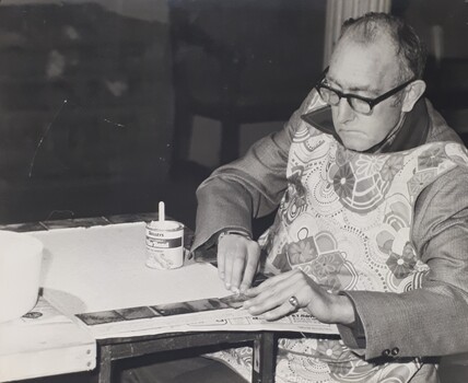 Seated man wearing apron as he adheres tiles to a board