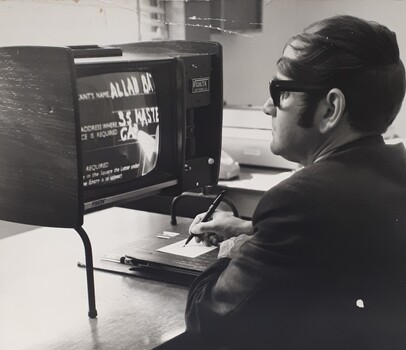 Seated man looking at a screen magnifier as he writes on paper, placed on to a board