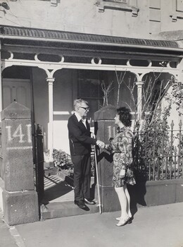 Man holding white cane, shakes hands with a female at his front gate