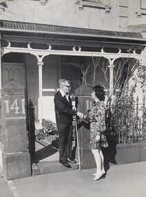 Man holding white cane, shakes hands with a female at his front gate