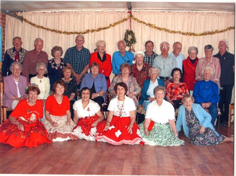 Large group of people sitting, standing and on floor looking towards camera, some dressed in square dance skirt