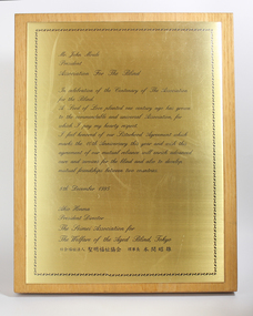 Gold coloured metal plate with cursive writing attached to wooden board
