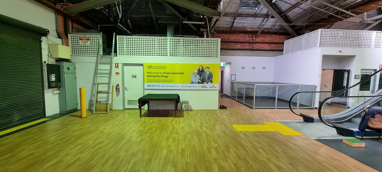 Yellow Vision Australia sign with blue writing on wall with enclosure and escalators to the right