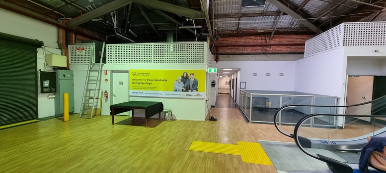 Yellow Vision Australia sign with blue writing on wall with enclosure and escalators to the right