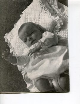 Baby on white bedding reaches one hand towards the sky