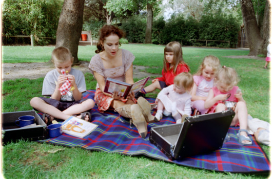 Sigrid and five children sit on picnic blanket as she reads a story, with two open suitcases beside them