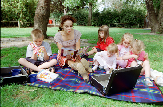 Sigrid and five children sit on picnic blanket as she reads a story, with two open suitcases beside them