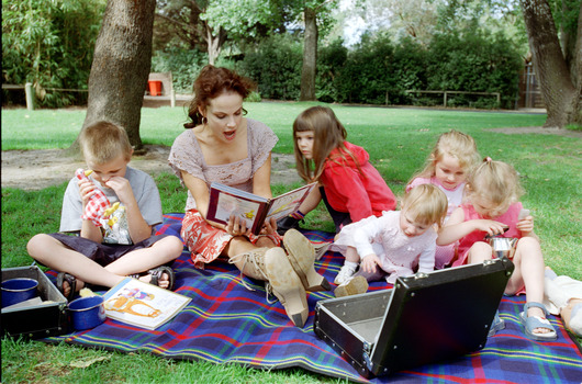 Sigrid and five children sit on picnic blanket as she reads a story, with two open suitcases beside them