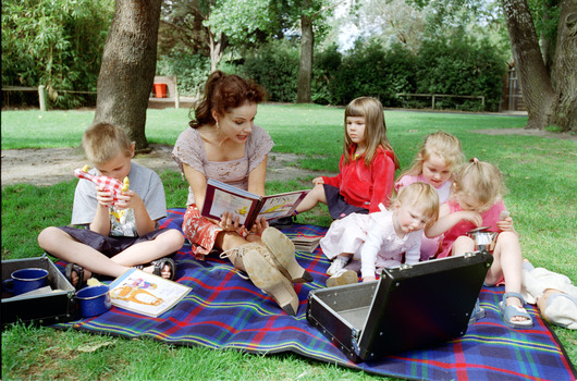 Sigrid and five children sit on picnic blanket as she reads a story, with two open suitcases beside them