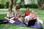Sigrid and five children sit on picnic blanket as she reads a story, with two open suitcases beside them