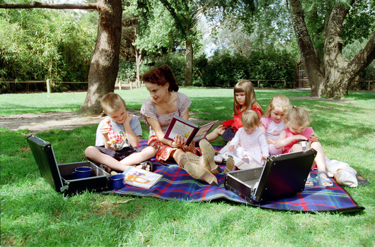 Sigrid and five children sit on picnic blanket as she reads a story, with two open suitcases beside them