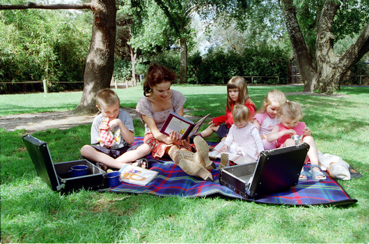 Sigrid and five children sit on picnic blanket as she reads a story, with two open suitcases beside them