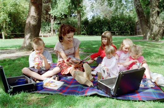 Sigrid and five children sit on picnic blanket as she reads a story, with two open suitcases beside them