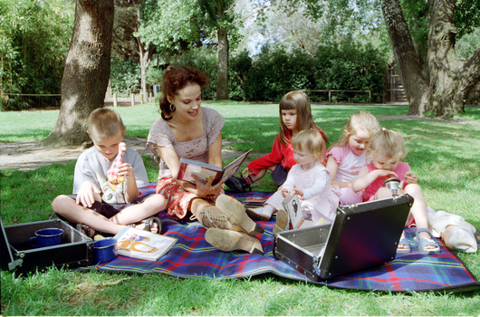 Sigrid and five children sit on picnic blanket as she reads a story, with two open suitcases beside them