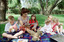 Sigrid and five children sit on picnic blanket as she reads a story, with two open suitcases beside them