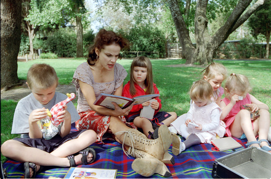 Sigrid and five children sit on picnic blanket as she reads a story, with two open suitcases beside them