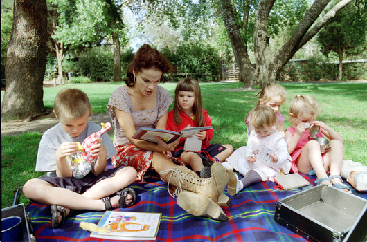 Sigrid and five children sit on picnic blanket as she reads a story, with two open suitcases beside them