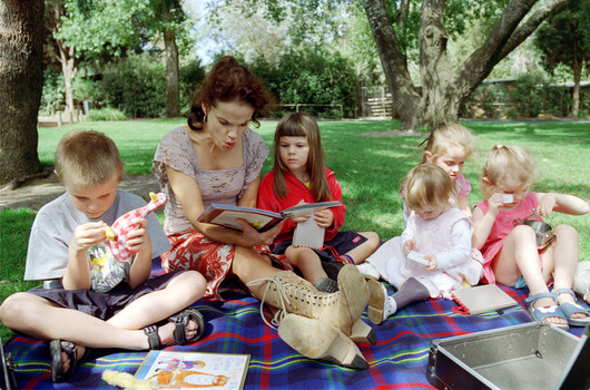 Sigrid and five children sit on picnic blanket as she reads a story, with two open suitcases beside them