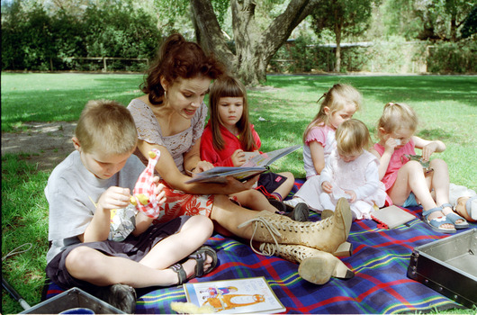 Sigrid and five children sit on picnic blanket as she reads a story, with two open suitcases beside them