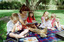 Sigrid and five children sit on picnic blanket as she reads a story, with two open suitcases beside them