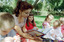 Sigrid and five children sit on picnic blanket as she reads a story, with two open suitcases beside them