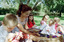 Sigrid and five children sit on picnic blanket as she reads a story, with two open suitcases beside them